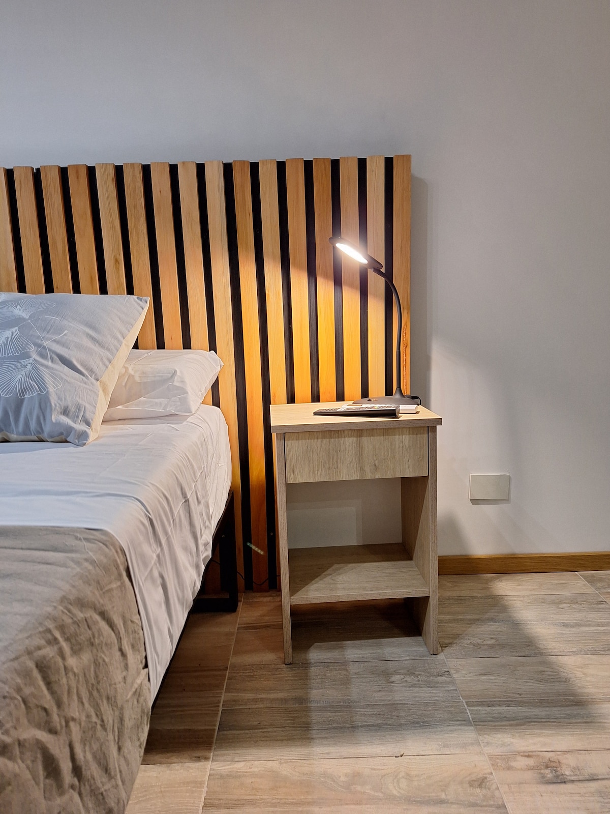 A well-appointed sleeping area featuring a spacious bed with a decorative headboard made of vertical wooden slats. A bedside table with a lamp and a small decorative item is positioned beside the bed, while natural light illuminates the warm wood-toned flooring.