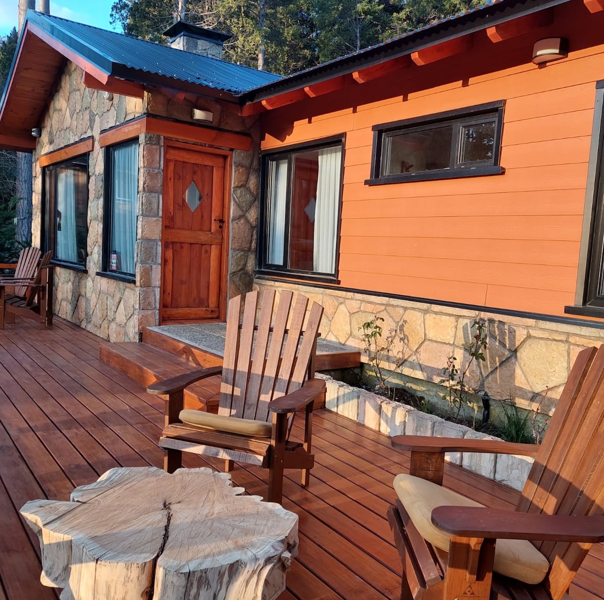 The exterior of the cabin is highlighted by natural stone and wooden accents, featuring a spacious deck with two wooden Adirondack chairs. Large windows allow natural light to brighten the inviting entrance, framed by a stone pathway and greenery.