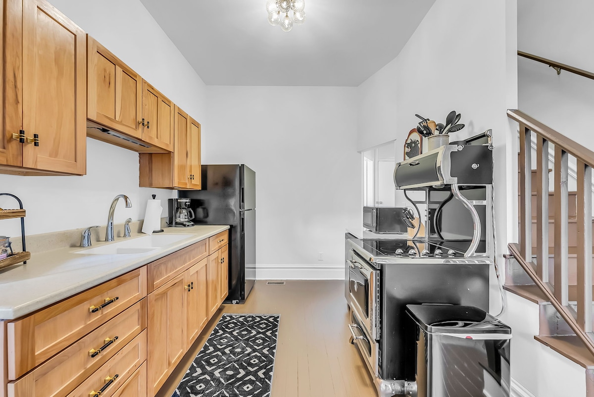 A well-equipped kitchen features wooden cabinetry and a spacious countertop. Stainless steel appliances are present, including a refrigerator and a commercial-style range. A dark-patterned rug adds contrast to the light wood floor, while ample natural light illuminates the space.