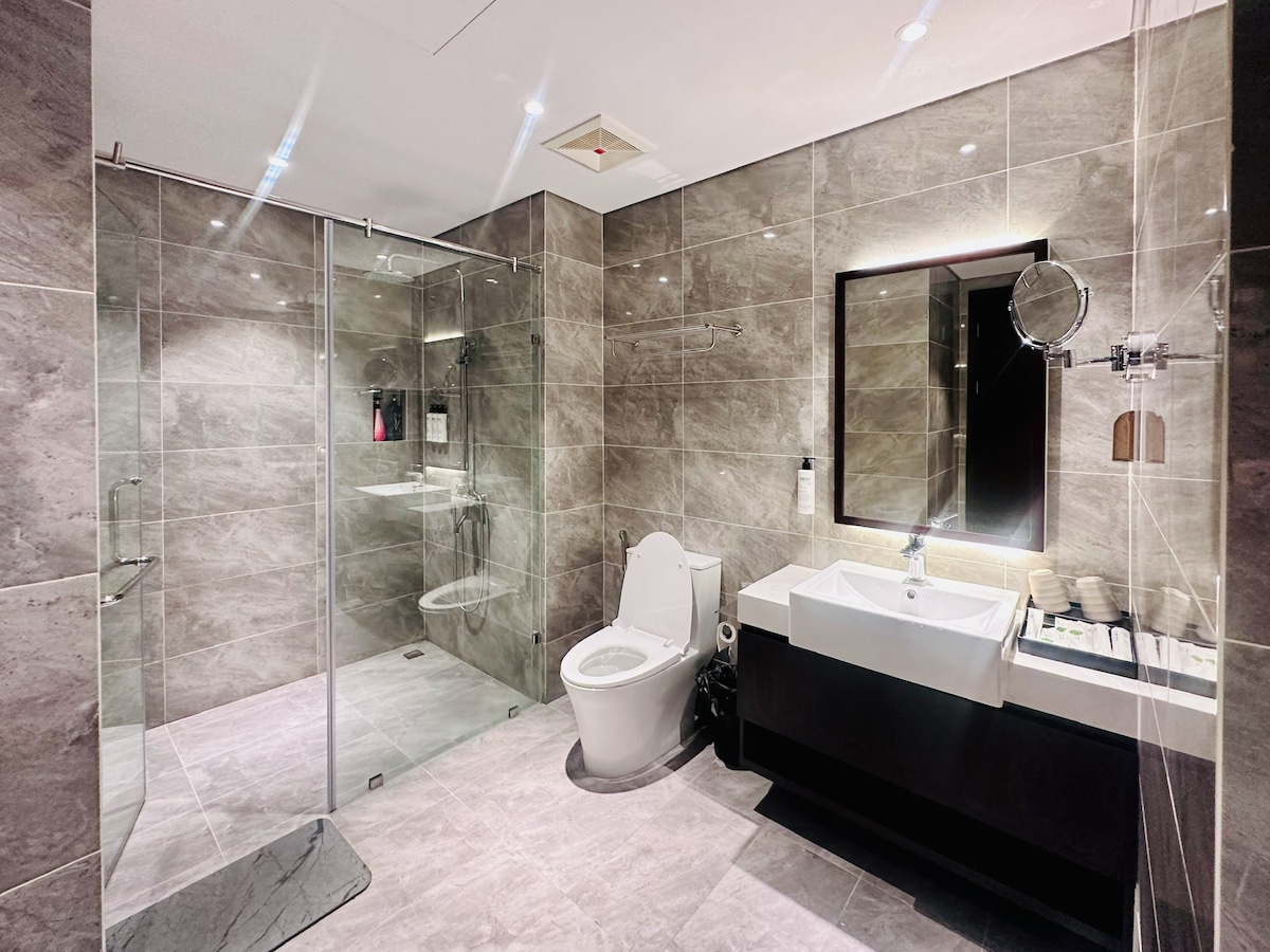 The bathroom features modern gray tiling and a spacious layout. A large glass shower enclosure is visible alongside a freestanding white sink with a dark wood cabinet beneath. Essential toiletries and towels are neatly arranged for guest convenience. A large mirror is mounted above the sink.