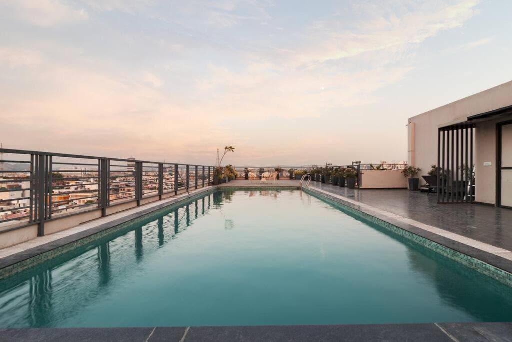 A rooftop pool stretches across the image, reflecting the pastel colors of the evening sky. Surrounding greenery and evening lights create a tranquil atmosphere, complemented by a railing that provides safety and scenic views of the city landscape.