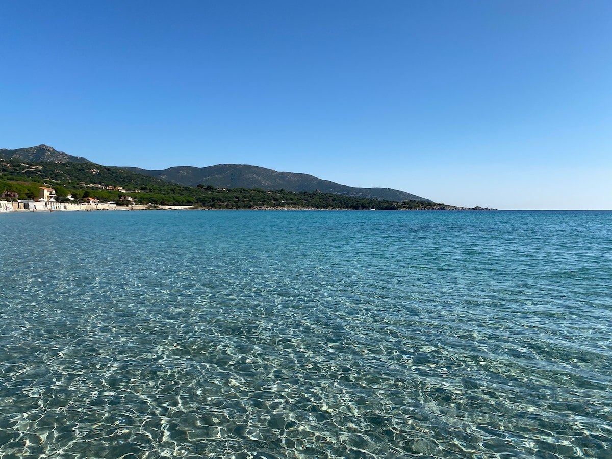 A clear and calm sea reflects the blue sky, with gentle waves lapping at the shore. In the background, hills and distant land highlight the serene coastline, inviting relaxation and exploring the natural beauty of the area.