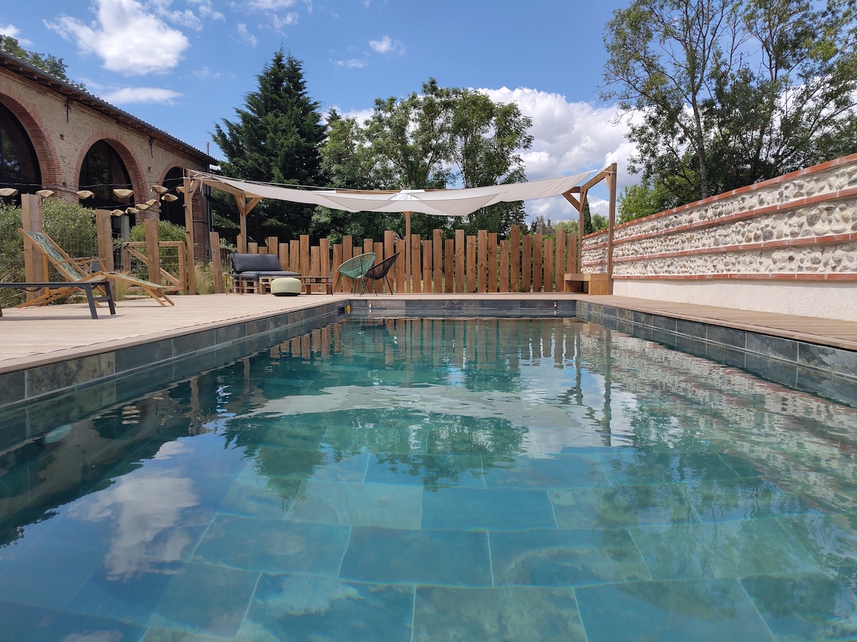 An inviting outdoor pool features clear, turquoise water, surrounded by wooden decking and shaded by fabric canopies. Wooden loungers are arranged nearby, with lush greenery and a rustic wall providing a scenic backdrop. The open sky above enhances the tranquil environment.
