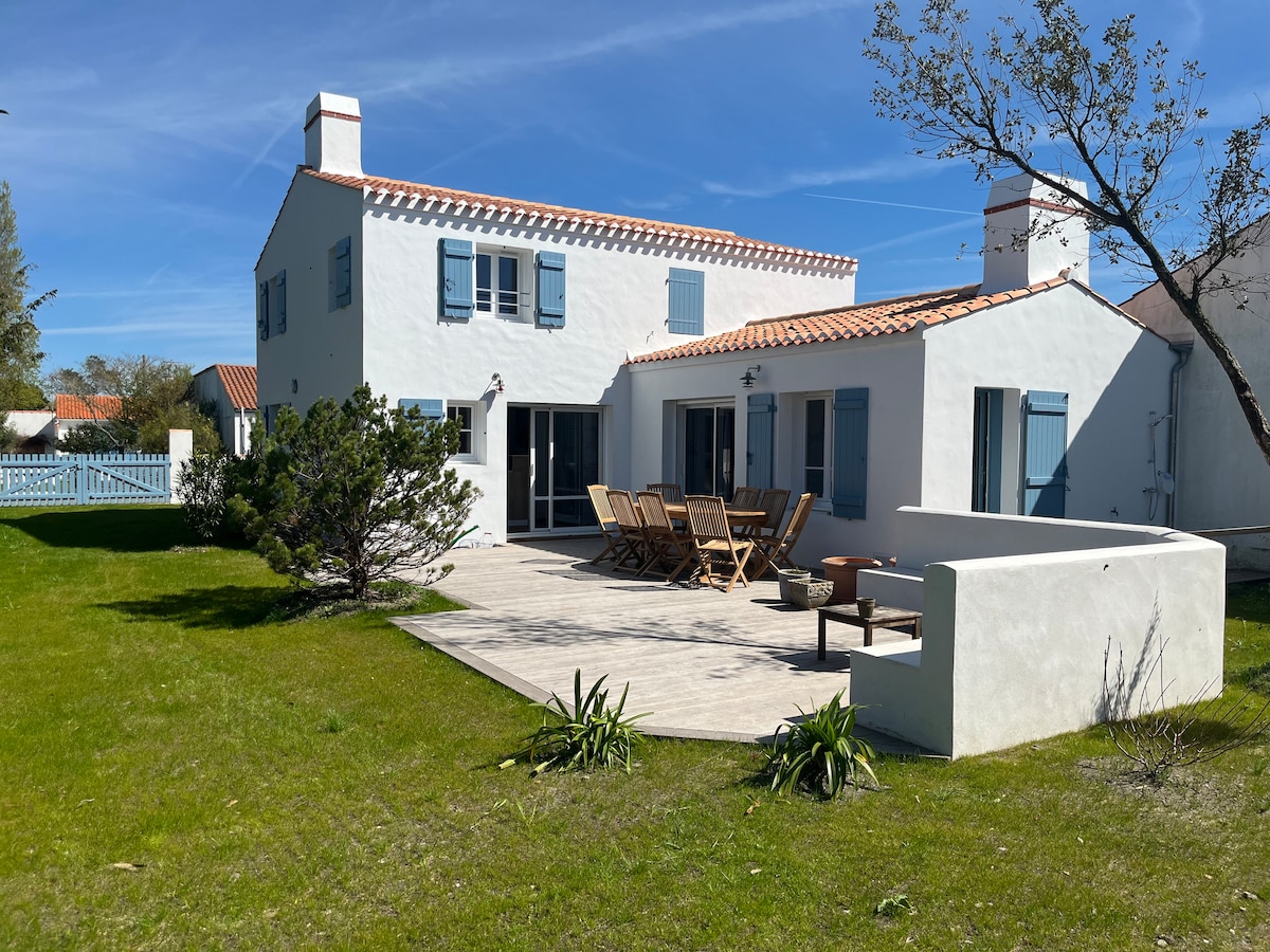 The exterior of the newly constructed house is shown, featuring a spacious garden with greenery and a large terrace equipped with a dining table and chairs. The house is characterized by white walls and blue shutters, with large glass doors connecting the indoors to the outdoors.