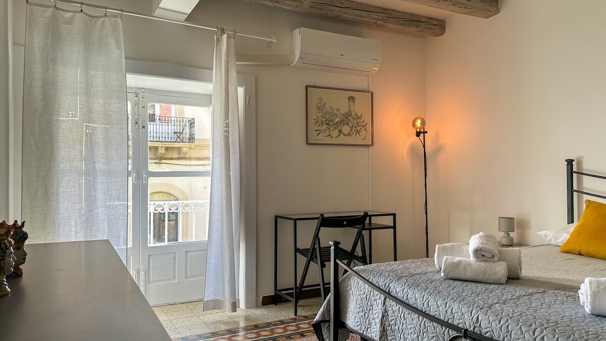 A cozy bedroom features a spacious bed adorned with fresh linens and decorative pillows. Natural light streams in through a balcony door, accentuated by sheer curtains. A small table and chair are positioned nearby, while exposed beams add a charming element to the room.