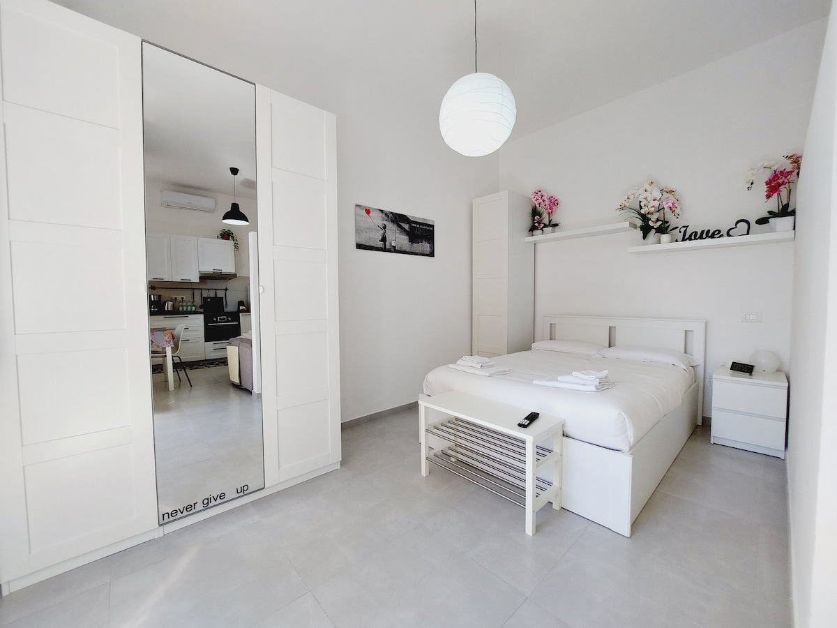 A bright and minimalistic bedroom features a spacious white bed with fresh linens. Decorative flowers are positioned on a shelf above, and a large mirror reflects the opposite wall, adding depth to the space. Soft lighting is provided by a pendant lamp.