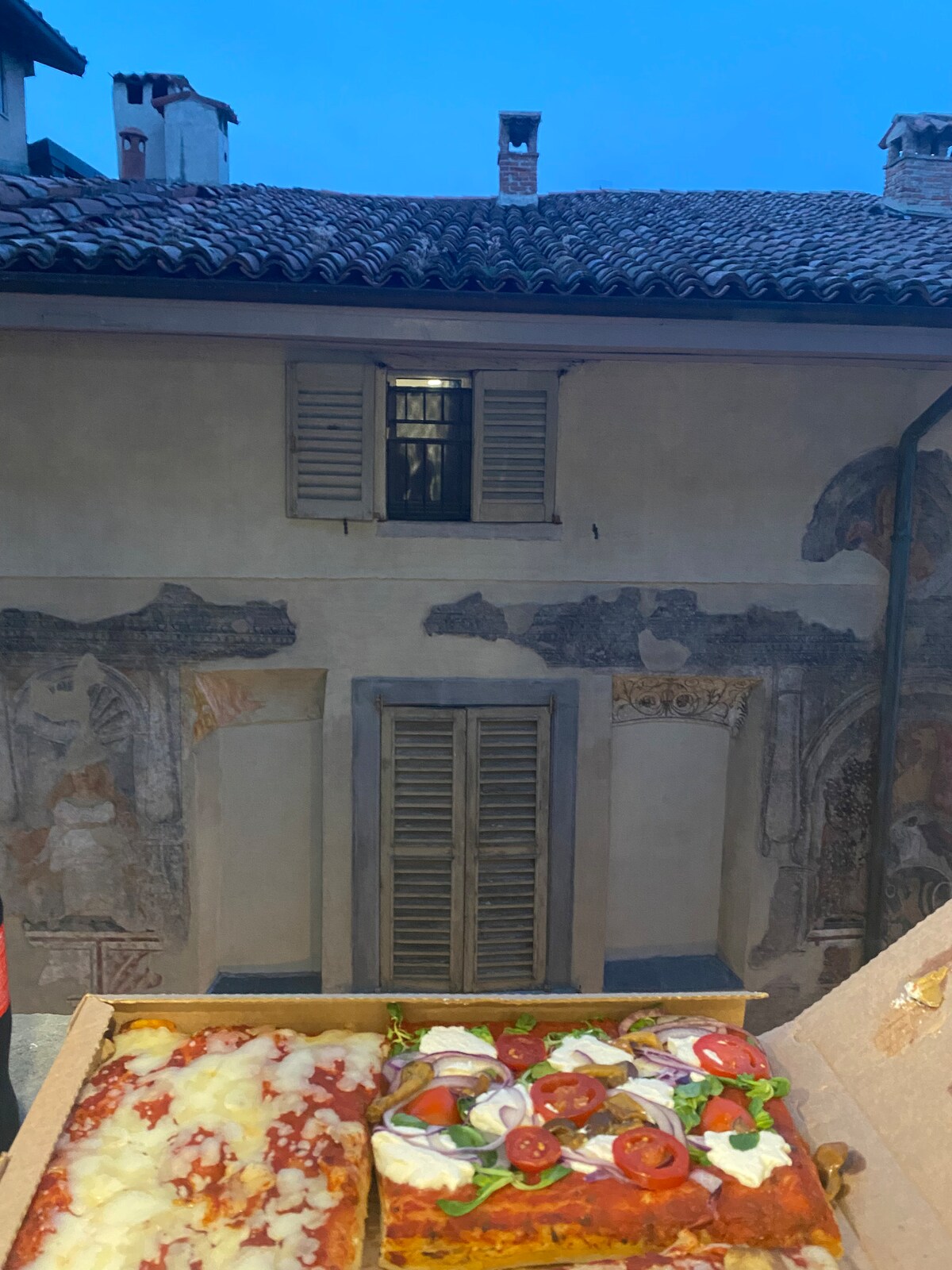 A historic building features faded frescoes and wooden shutters, reflecting the charm of the area. The image captures a glimpse of the building’s architecture, framed by an evening sky. A pizza box in the foreground hints at a local culinary experience.