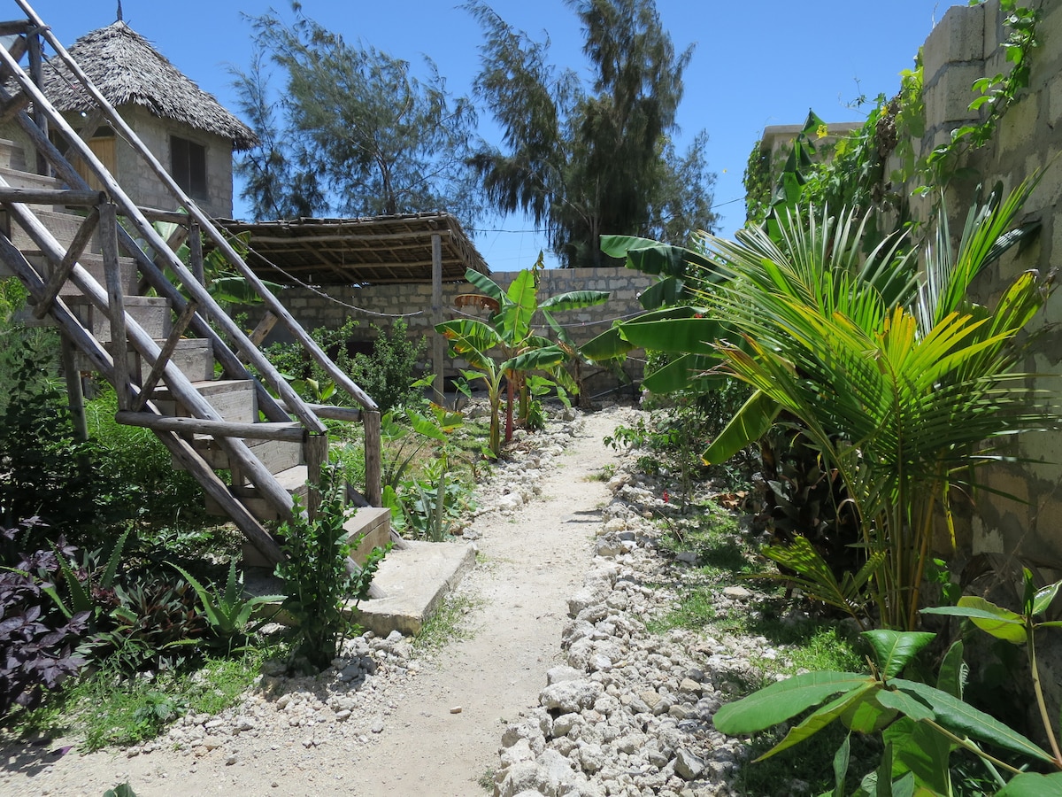 A winding pathway, bordered by lush greenery, leads through a garden area. Tropical plants, including banana and palm trees, are visible alongside stone arrangements. A wooden staircase is situated to the left, providing access to an elevated space, while blue skies and sunlight enhance the outdoor setting.
