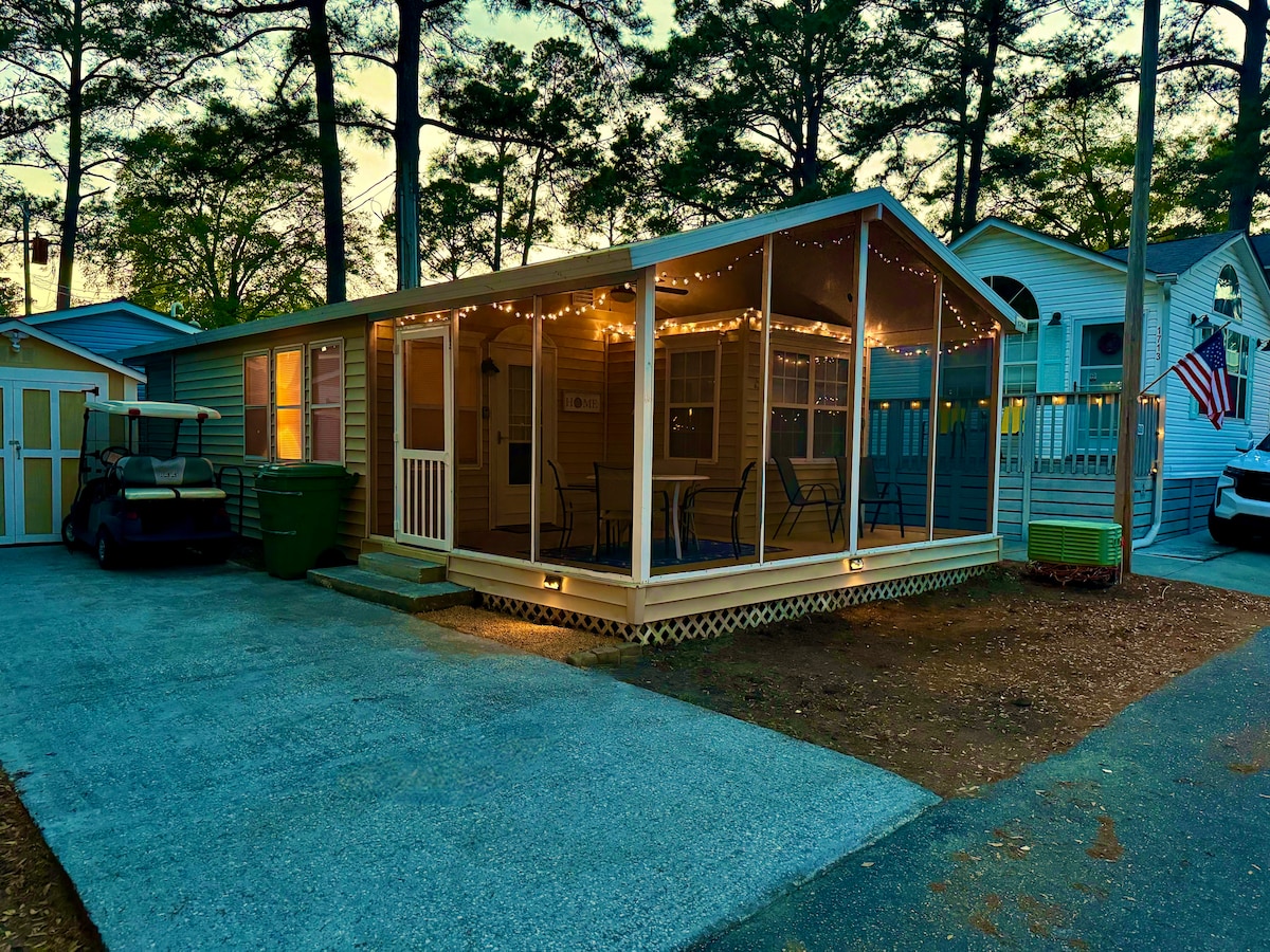 The exterior view of the cottage is displayed, featuring a screened porch adorned with string lights. A golf cart is parked nearby, alongside a driveway with gravel. The entry is framed by a white door with a small window, inviting guests into the home.