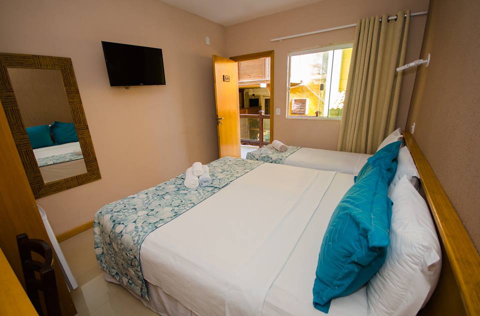 A well-appointed room features two beds, one with a patterned coverlet and the other topped with a solid white duvet. A mirror and a television are mounted on the walls, while natural light enters through a window adorned with light-colored drapes.