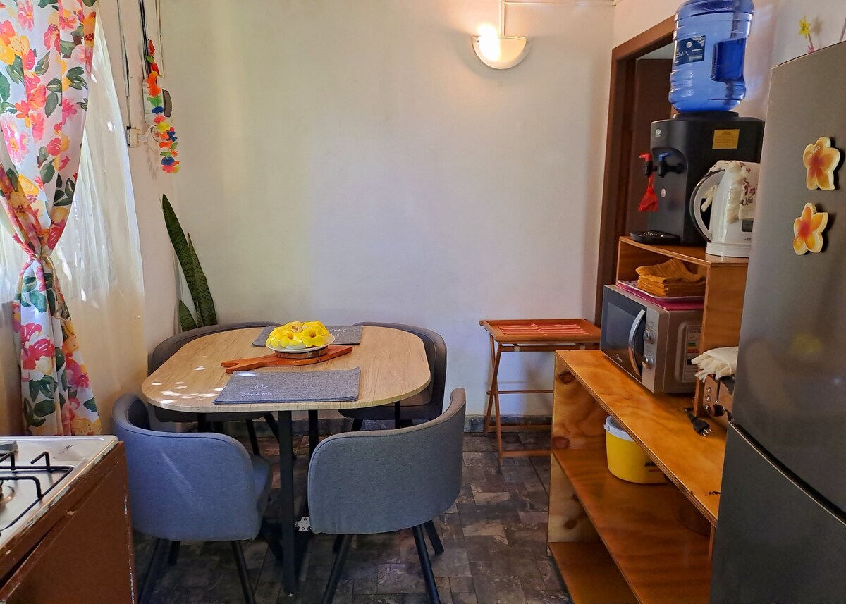 A cozy kitchen space is visible, featuring a round dining table surrounded by four dark chairs. Bright floral curtains frame a window, while a small shelf holds a microwave and other kitchen essentials. Natural light adds warmth to the overall atmosphere.