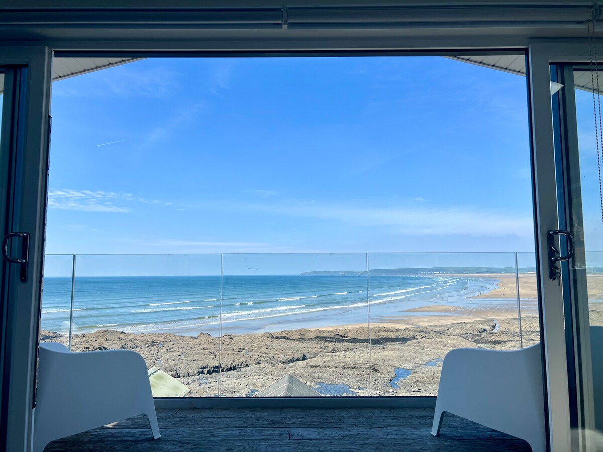 A clear blue sky is framed by expansive glass doors, revealing a panoramic view of the beach and ocean. Gentle waves roll onto the shore, and two modern chairs are positioned on a balcony, offering a relaxing spot to enjoy the scenic coastal landscape.