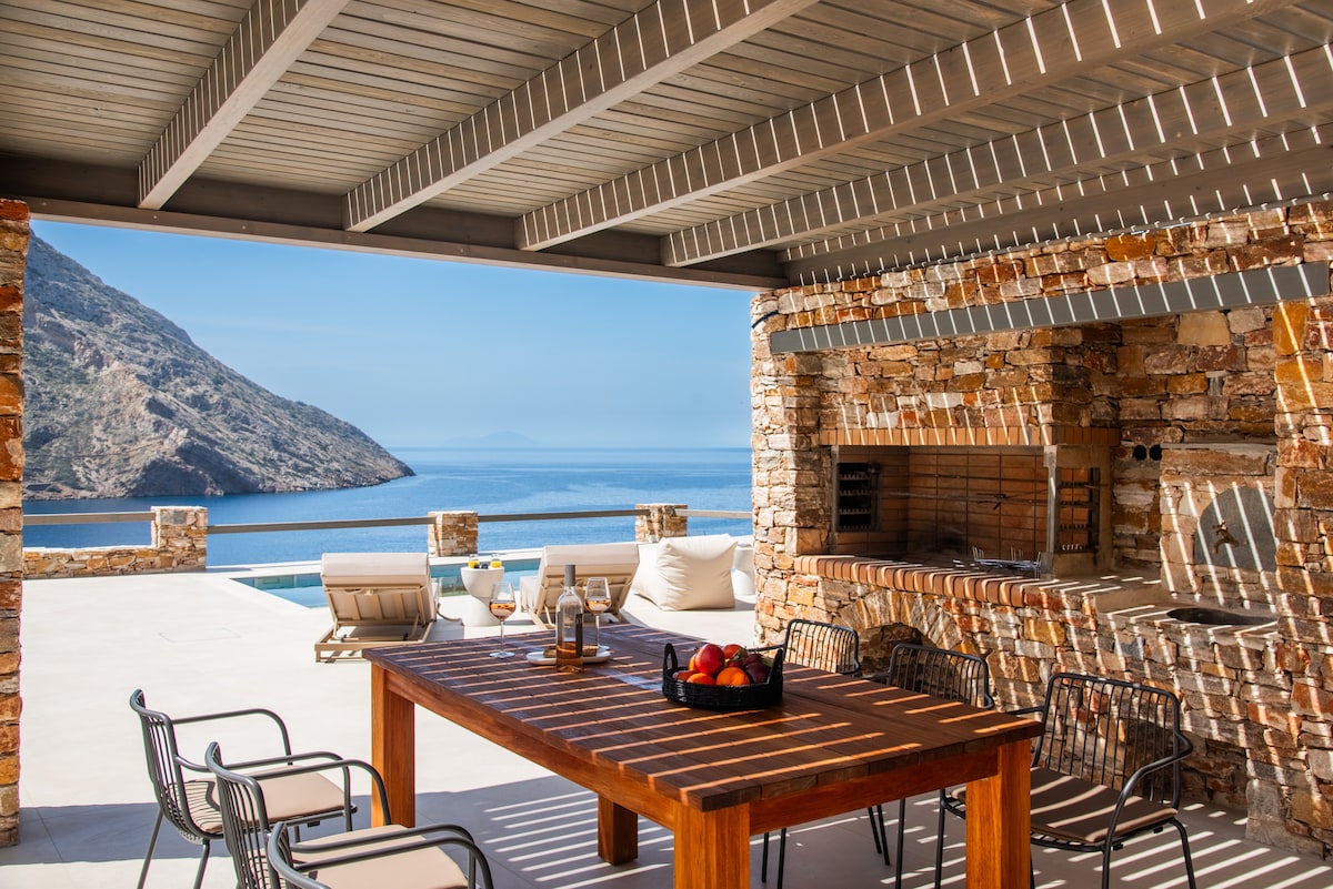 An outdoor dining area features a rustic wooden table surrounded by metal chairs. A stone barbecue grill stands nearby, with a panoramic view of the ocean and mountains in the background. Natural light filters through the overhead beams, creating varying patterns on the floor.