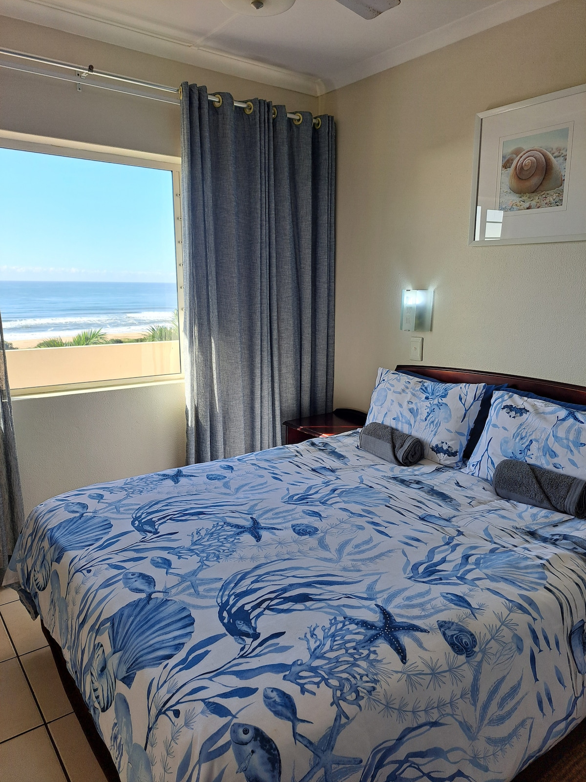 A light-filled bedroom features a spacious bed adorned with blue marine-themed bedding. Large windows offer a view of the beach, while sheer curtains frame the space. Nightstands on each side of the bed hold decorative lamps, enhancing the room's functionality.
