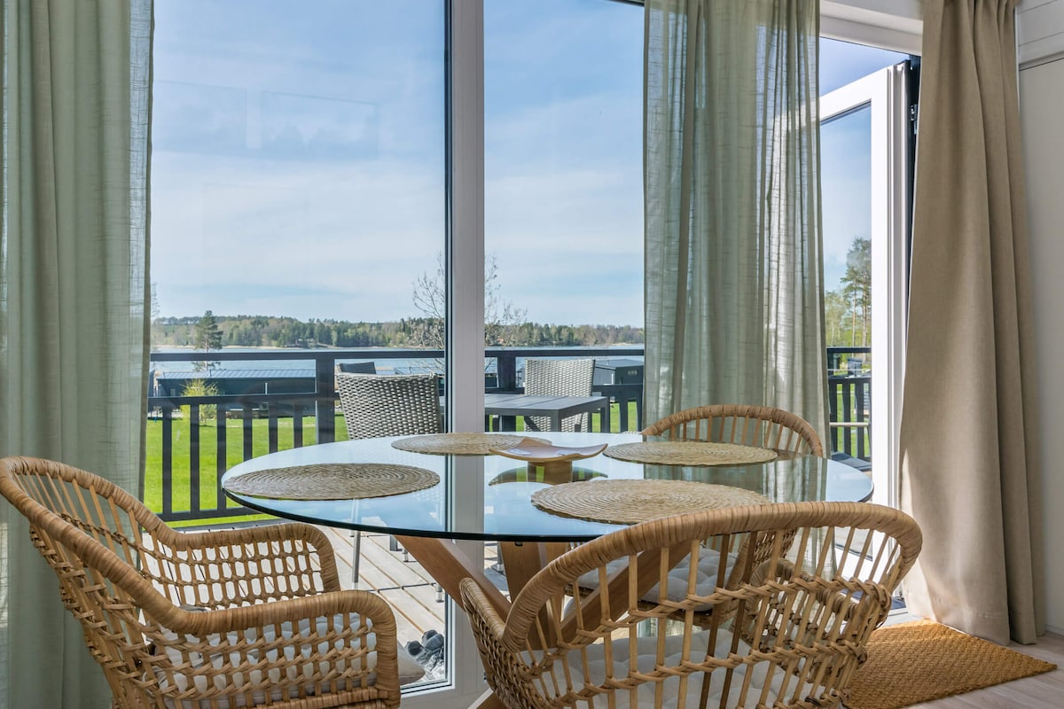 An inviting dining area is captured with two woven chairs surrounding a round glass table. Floor-to-ceiling windows allow for abundant natural light and showcase serene views of the greenery and water beyond. Light curtains provide a soft contrast to the bright setting.