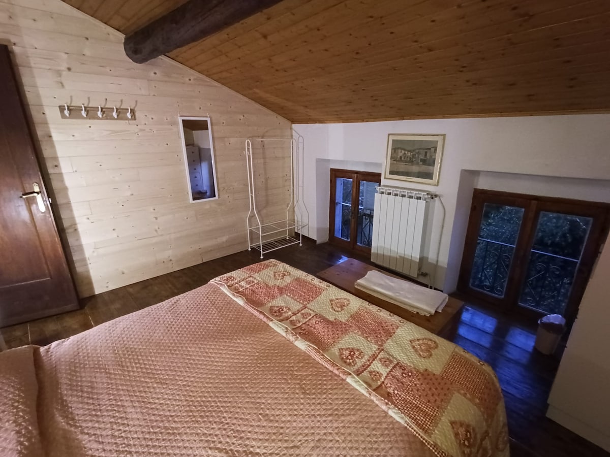 A cozy bedroom is featured with a sloped wooden ceiling. Natural light enters through two small windows. A spacious bed adorned with a patterned blanket is positioned prominently, while a minimalistic clothing rack and a mirror add functional elements to the room.