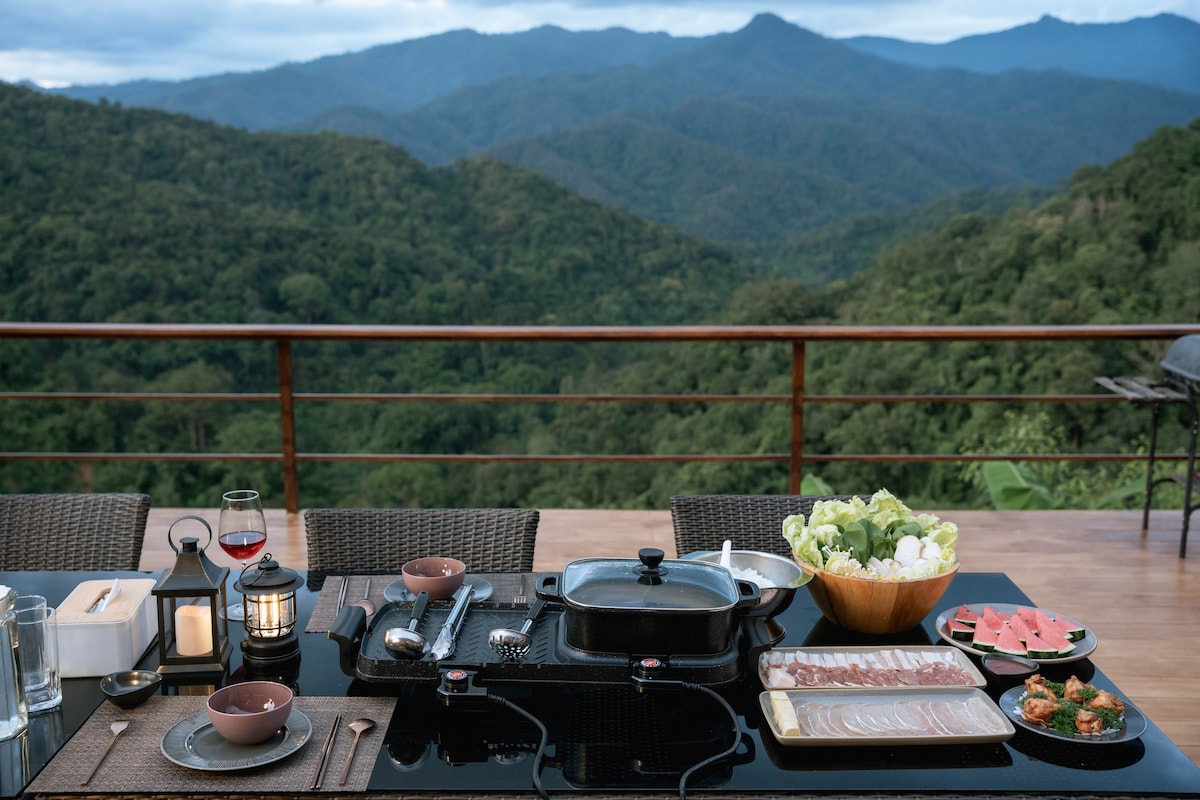 A dining table is set with various dishes and utensils, overlooking a lush mountain landscape. A portable grill and plates with fresh ingredients are displayed, accompanied by a candle and drinks. The expansive view complements the dining experience, providing a serene backdrop.