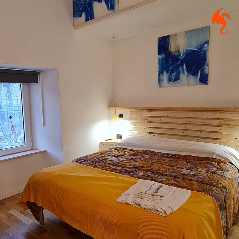 A spacious bedroom features a wooden bed frame with a plush bedspread in warm tones. An abstract artwork adorns the wall above the bed. Natural light filters through the window, creating a calm and inviting space.