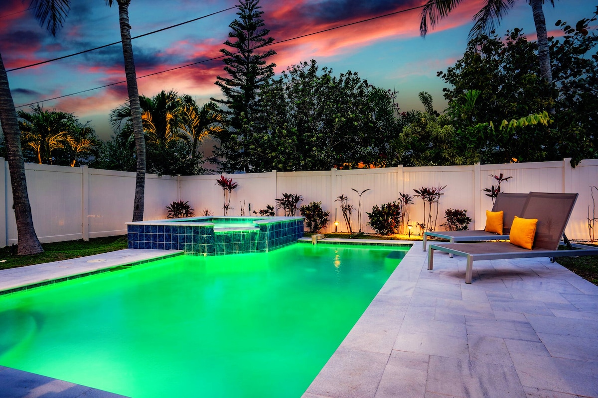 A serene outdoor space is highlighted by a heated pool and spa, featuring glowing turquoise water. The area is bordered by lush greenery and a white privacy fence, with comfortable lounge chairs positioned nearby. The sky is painted in hues of pink and purple, adding to the tranquil ambiance.
