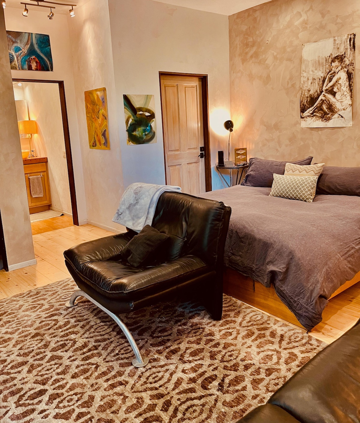 A cozy interior features a spacious bed with grey bedding, complemented by decorative pillows. A plush black chair, positioned on a patterned rug, offers seating. Light is softly diffused through the room, highlighting artistic wall decor and access to an adjoining bathroom area.