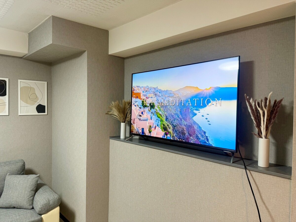 A modern living area is featured, showcasing a large flat-screen TV mounted on a minimalist wall. A cozy gray sofa is positioned nearby, complemented by framed artwork on the wall. Natural decorative elements are placed on the shelf beneath the TV.