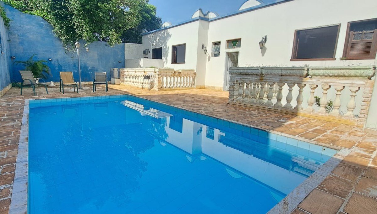 A refreshing pool is situated in a sunlit courtyard, surrounded by a tiled deck. Lounge chairs are positioned nearby, and decorative balustrades frame the space, complementing the white walls and vibrant blue accents of the background.