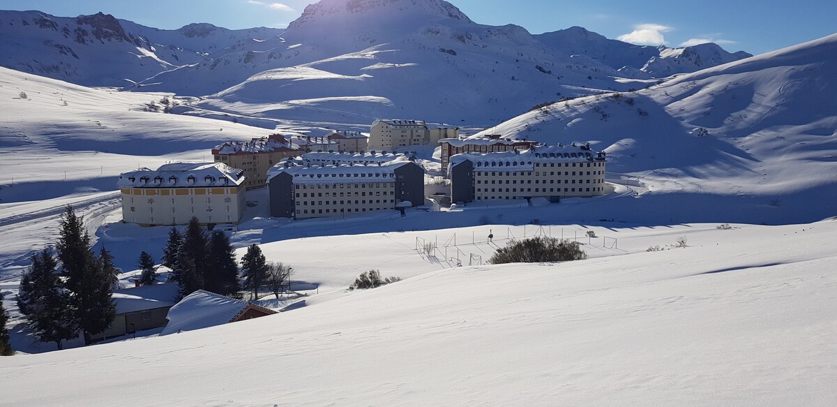 A winter landscape captures a cluster of buildings nestled among snow-covered hills. The structures, blanketed in white, display uniform facades. Snow-draped trees are positioned in the foreground, and distant mountains stretch across the horizon under a clear blue sky.