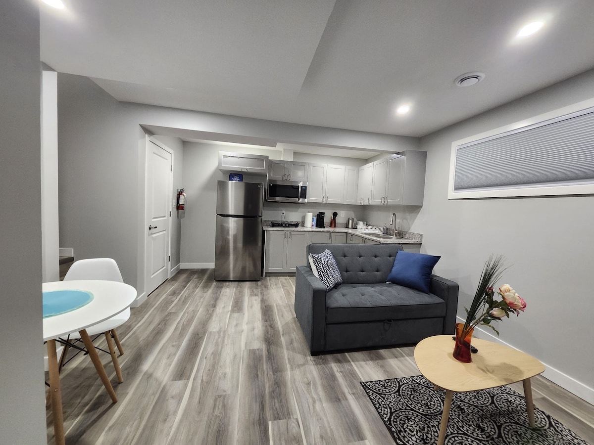 The modern basement suite features a cozy sitting area with a gray sofa and a small coffee table adorned with flowers. A dining table with white chairs is positioned nearby. Stainless steel appliances are visible in the kitchen area, enhancing the clean, open layout.