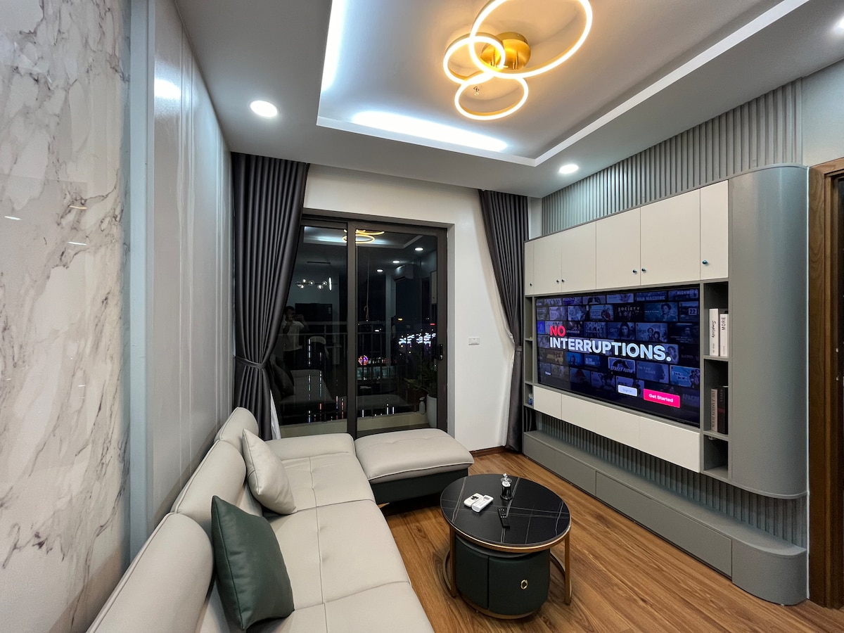 A modern living area is highlighted by a spacious sectional sofa and a sleek coffee table. Large windows provide natural light and city views, while a flat-screen TV mounted on cabinetry adds a contemporary touch. The room features elegant lighting and floor-to-ceiling curtains.