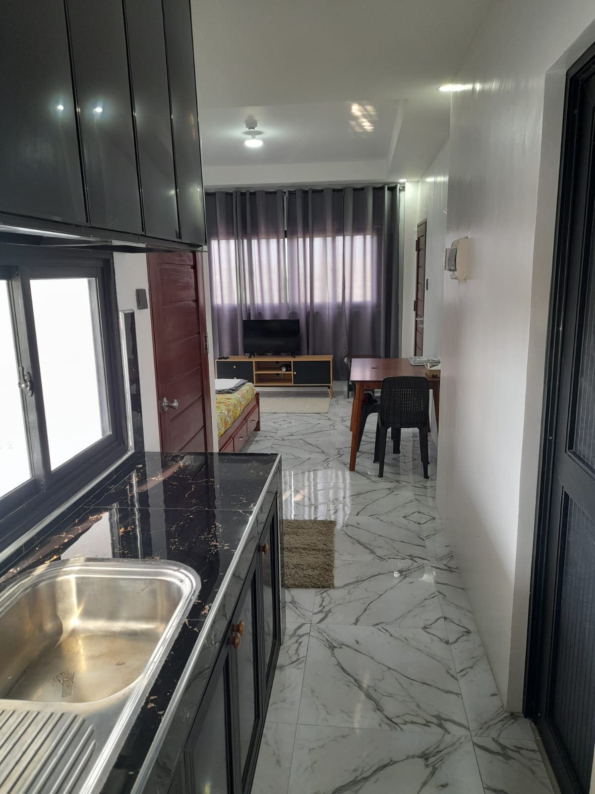 A compact studio kitchen features a stainless steel sink set against sleek black cabinets. The space transitions into a living area highlighted by a window adorned with long gray curtains. A wooden dining table and chair occupy the room, alongside a small television unit.