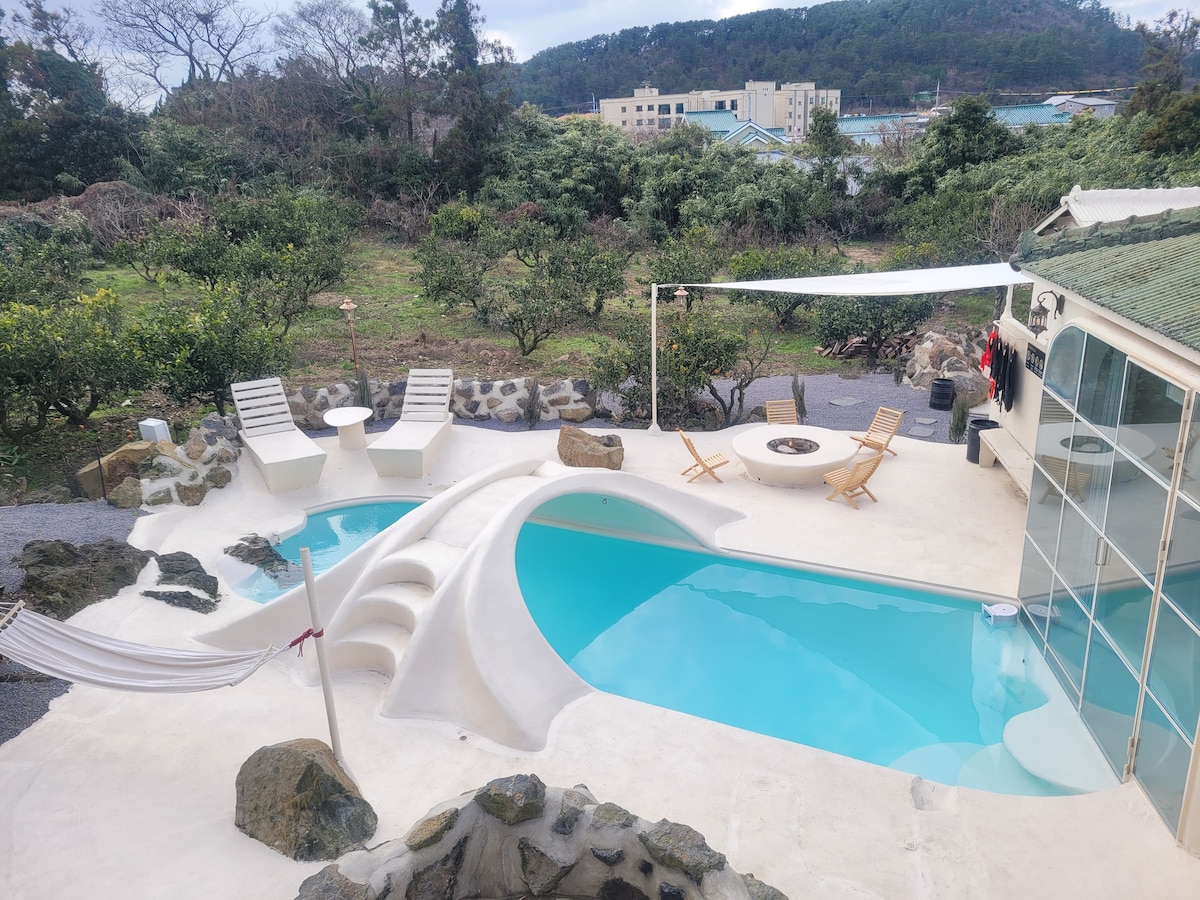 An outdoor area features a unique-shaped pool and a separate hot tub, surrounded by natural stone accents. Two sun loungers and a round seating area with a fire pit create spaces for relaxation. A hammock is positioned for additional comfort amidst a scenic backdrop.
