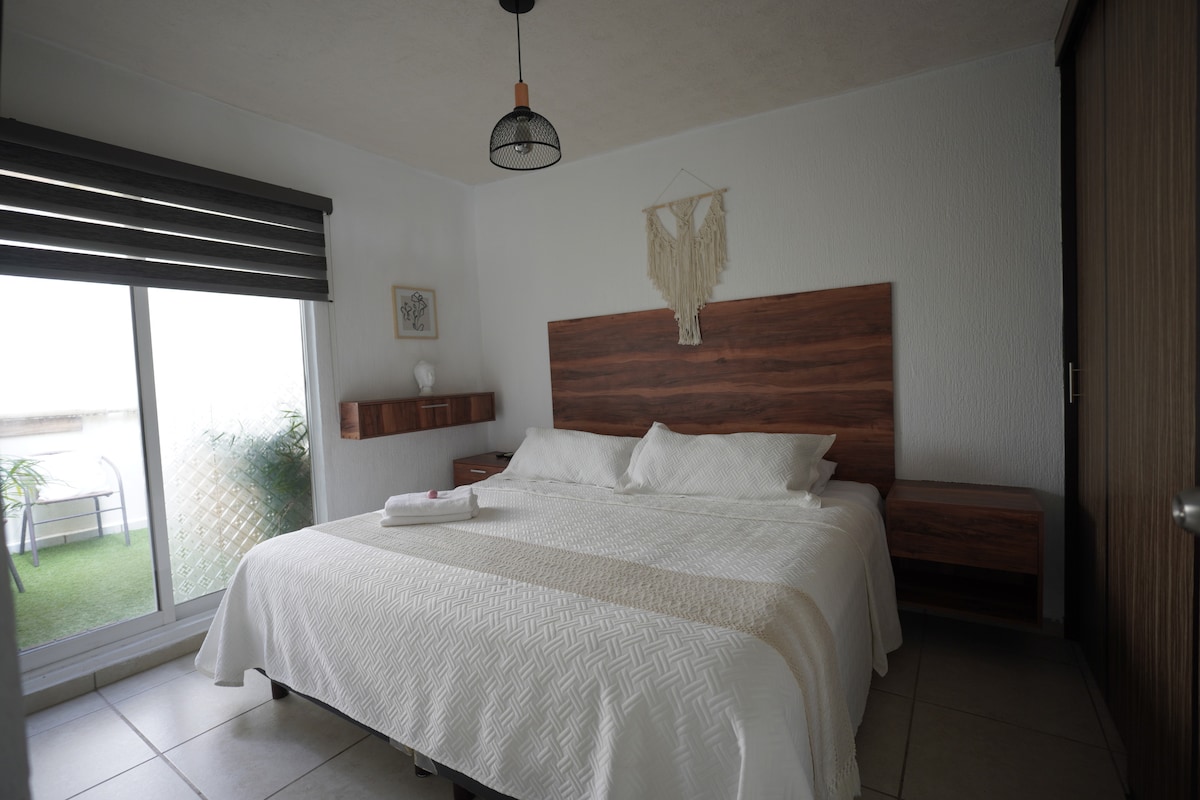 The room features a spacious bed with a neutral-colored quilt, complemented by two wooden nightstands. Natural light enters through the large sliding glass door, which opens to a small balcony. Decorative elements include a hanging wall art piece and a subtle indoor plant.
