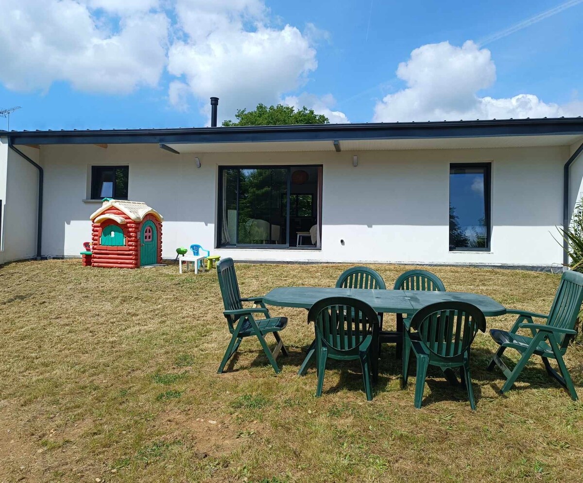 A spacious outdoor area features a green lawn with a six-seat dining table surrounded by matching chairs. A colorful playhouse is situated nearby, offering a playful element. The exterior wall of the house is white, with large glass doors allowing indoor visibility.