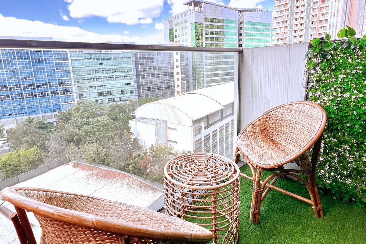 A relaxing balcony space features two wicker chairs and a small round table, surrounded by greenery and a scenic city view. The combination of natural textures and greenery enhances the inviting atmosphere, providing an ideal spot for enjoying fresh air.