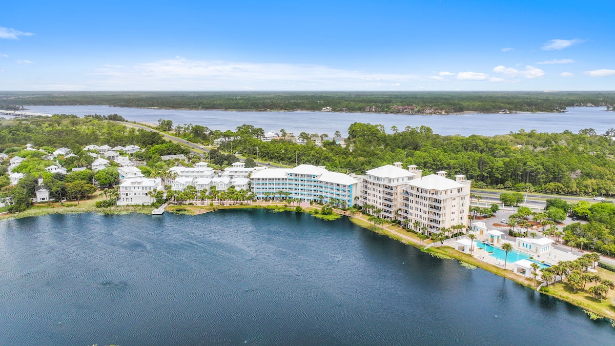 An aerial view captures the Carillon Beach community, showcasing a collection of white buildings along the serene lake's edge. Lush greenery surrounds the area, while the expansive water reflects the sky above. The image illustrates a peaceful setting, ideal for relaxation and outdoor activities.