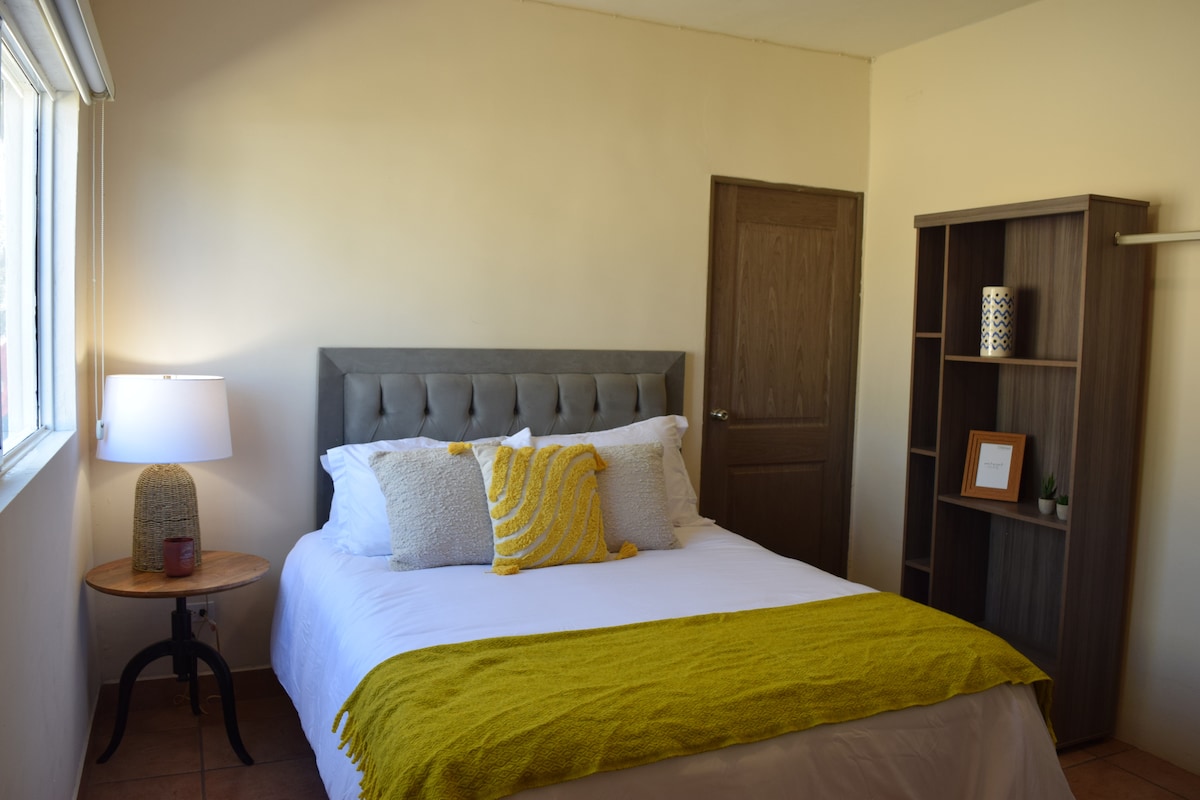 A cozy bedroom showcases a spacious bed dressed in white linens and complemented by a vibrant yellow throw. A bedside lamp emits soft light beside a small round table. A wooden bookshelf stands against the wall, adding functional elegance to the room.