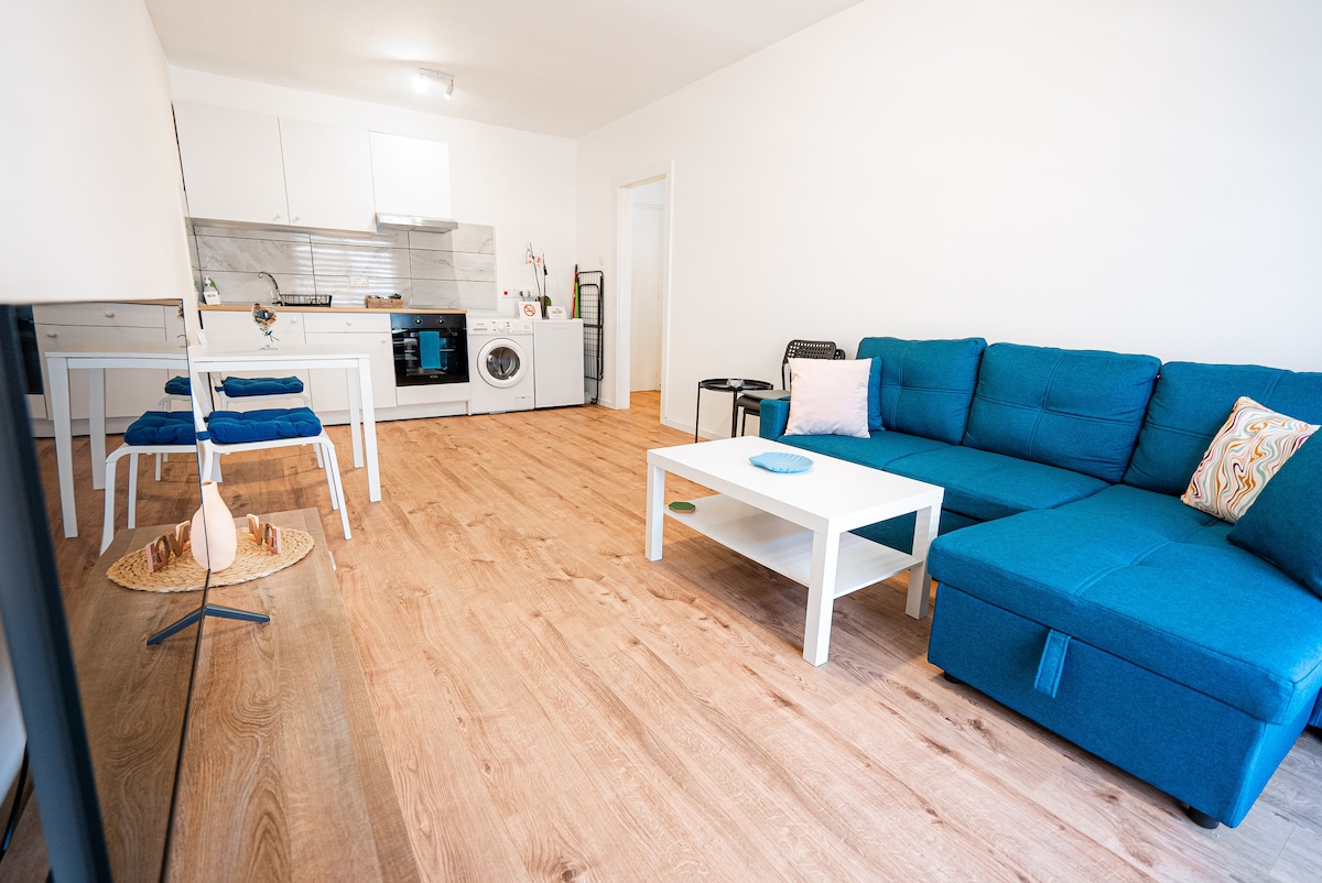A bright living area features a spacious blue sectional sofa and a white coffee table. A dining table with two chairs is positioned nearby. The open kitchen is visible, showcasing modern appliances and cabinetry. Light wood flooring complements the room's neutral color palette.