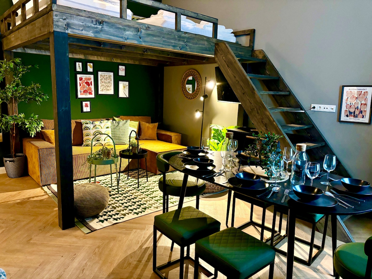 The space features a lofted sleeping area above a versatile living area with a couch and seating. A dining table for seven is set nearby, with black plates and glassware arranged. A staircase leads to the upper level, while greenery adds a touch of nature.