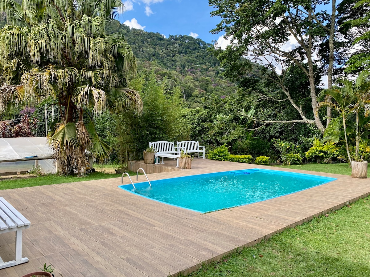 A serene outdoor area features a refreshing swimming pool surrounded by a wooden deck. Lush greenery and trees frame the space, providing a peaceful backdrop. Two white benches are positioned to enjoy the view, enhancing the welcoming atmosphere of the garden.