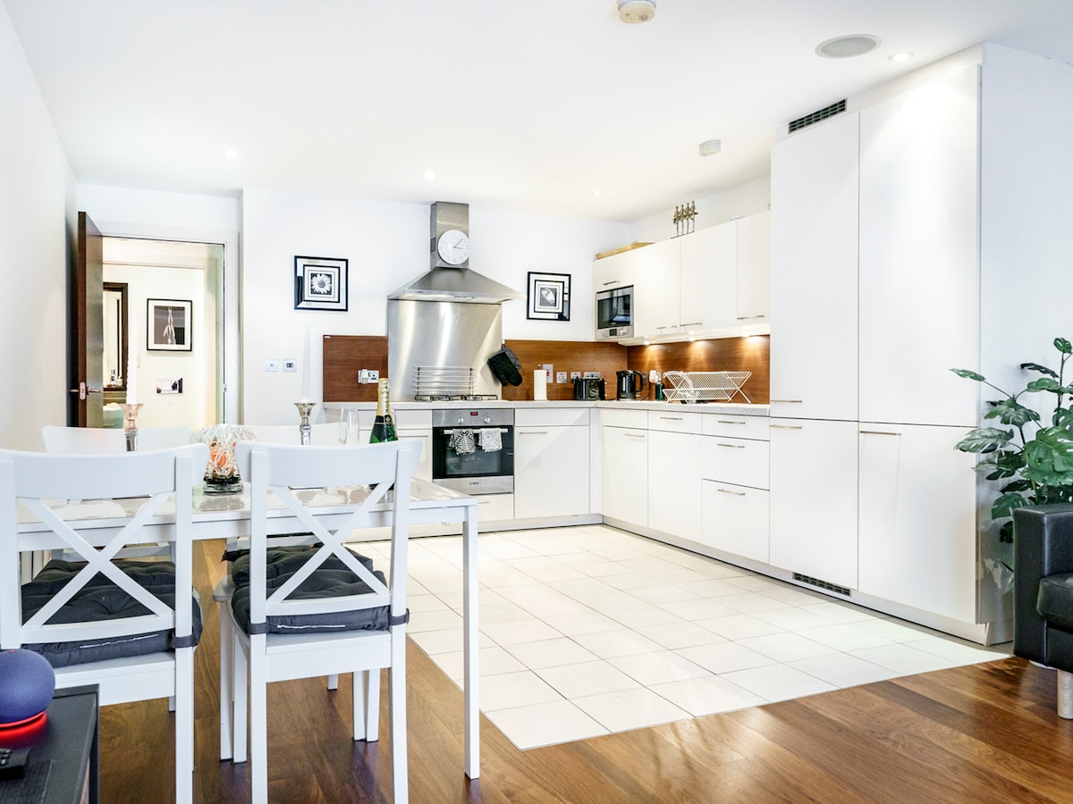 The kitchen features modern white cabinetry and stainless steel appliances, including an oven and extractor hood. A dining table with four chairs is positioned nearby, with a small plant adding greenery to the space. Sleek tile flooring enhances the contemporary atmosphere.