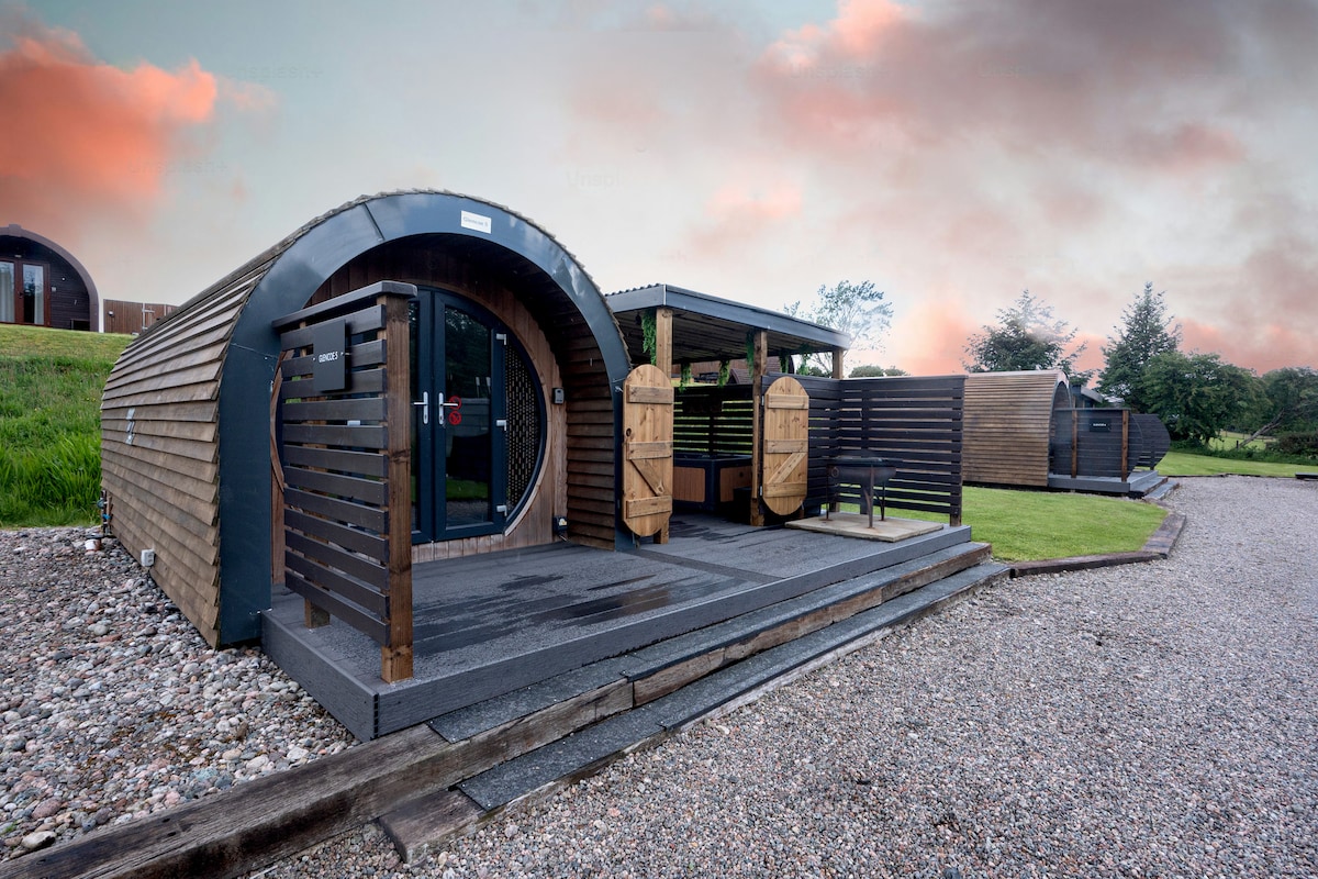 A cozy glamping pod is shown with a rounded wooden exterior and welcoming double doors. The private deck area features wooden barriers, enhancing privacy. Accessible pathways lead to a covered seating area, with lush green grass and a stone pathway visible in the surrounding space.