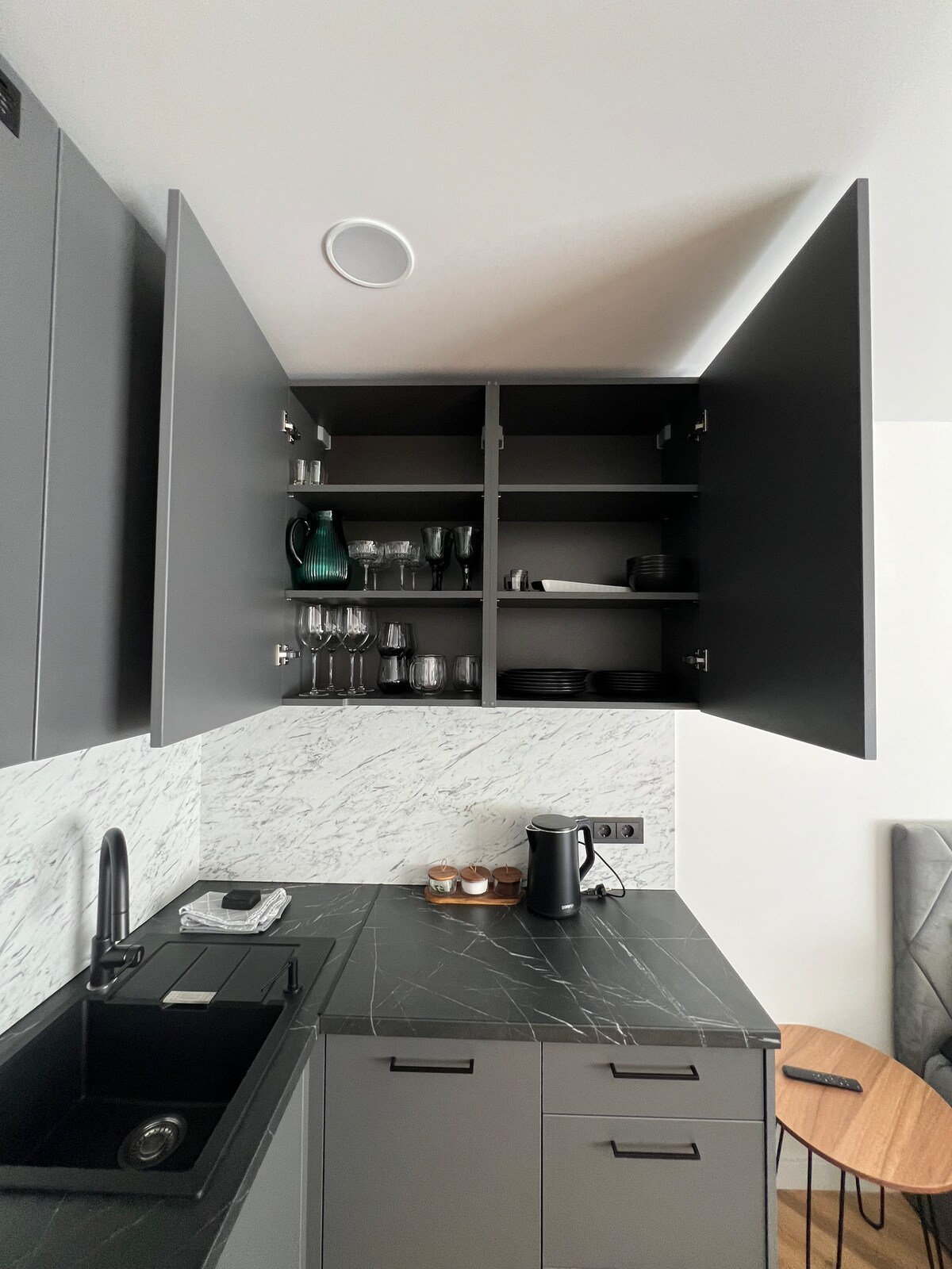 A modern kitchen features sleek grey cabinets with open doors revealing glassware and functional items. A minimalist black sink is visible, along with a small coffee maker on the countertop. Textured stone and wood elements contribute to the contemporary design.