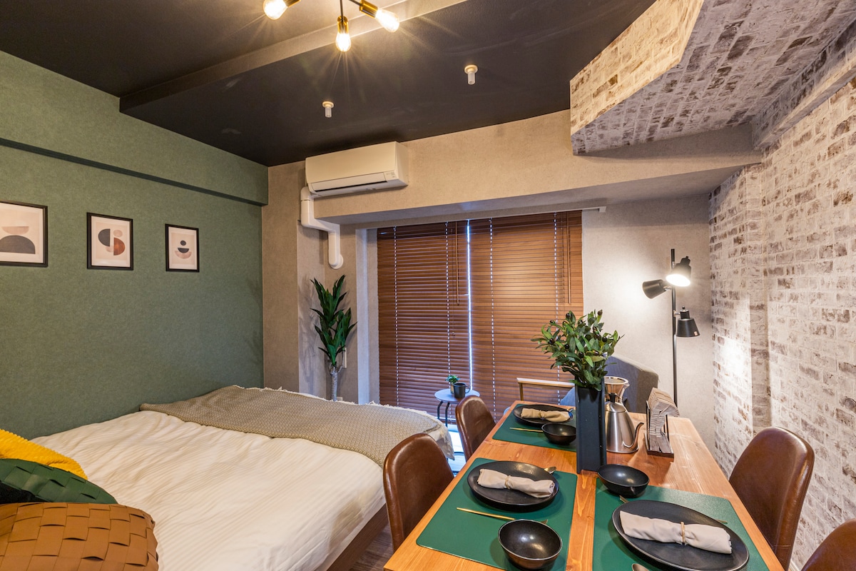 A stylish room showcases a cozy sleeping area with a spacious bed covered in soft linens. A dining table adorned with plates and cutlery is positioned beside the bed. Natural light filters through wooden blinds, highlighting a green plant and framed wall art.