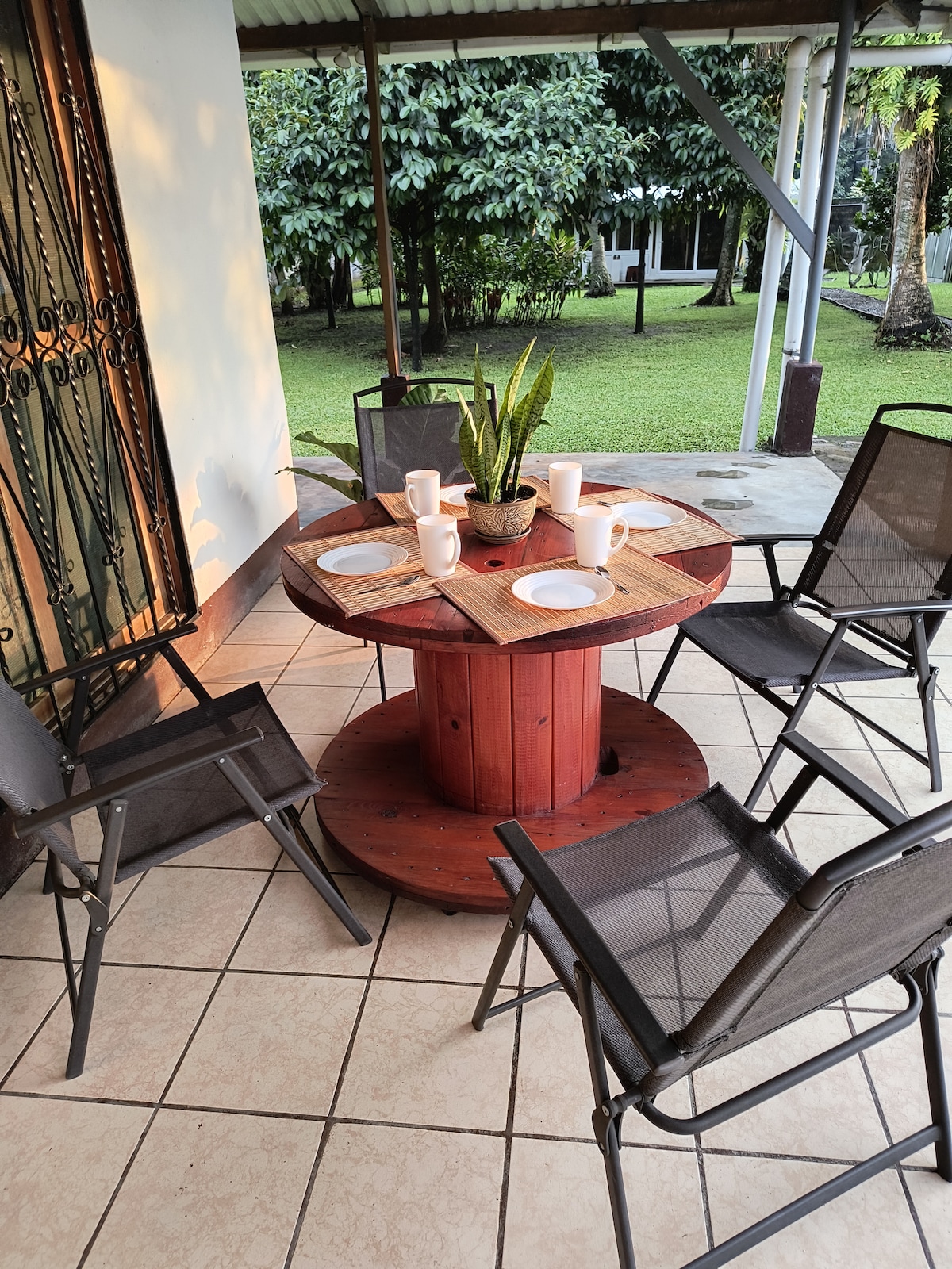 An outdoor dining area is presented with a circular wooden table, surrounded by six metal-framed chairs. A small plant sits in the center of the table, and white plates and mugs are arranged for use. Lush greenery surrounds the space, contributing to a relaxed atmosphere.