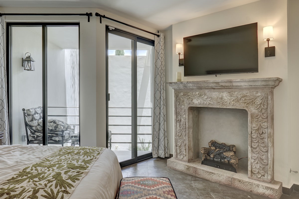 A serene bedroom features a spacious bed adorned with a decorative blanket. A stone fireplace adds a classic touch, complemented by a flat-screen TV mounted above it. Large glass doors lead to a private outdoor seating area, allowing natural light to fill the room.