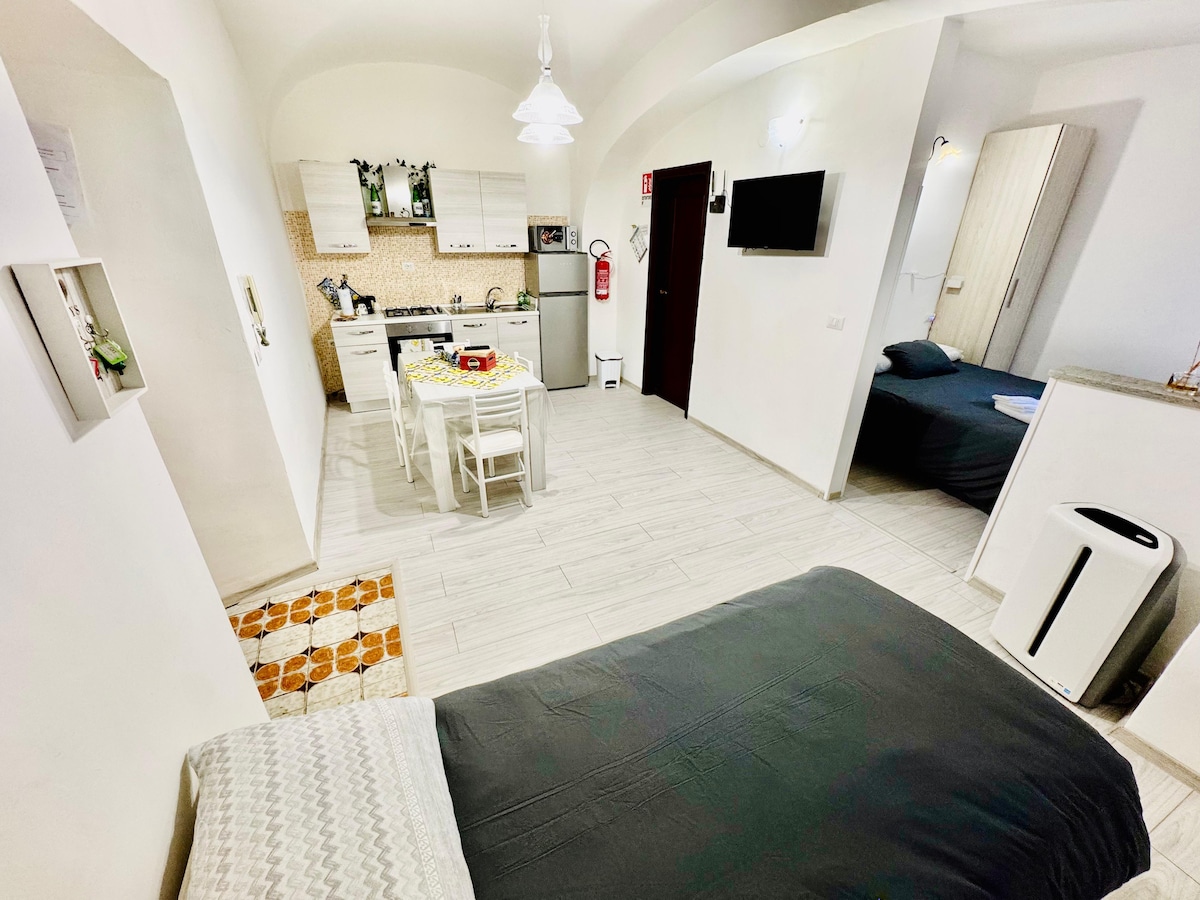 A spacious living area is presented, featuring a well-equipped kitchen with light cabinetry. A dining table with white chairs is visible, along with a cozy sleeping area that includes a dark bedspread. Soft lighting and a partial view of additional furnishings enhance the ambiance.