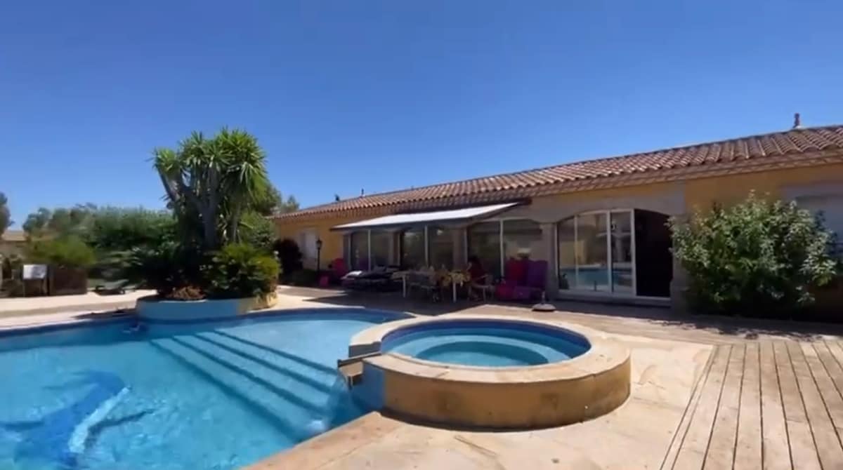 The outdoor area features a spacious swimming pool complemented by a separate circular whirlpool. Surrounding the pool are sun loungers and a shaded seating area. The villa's large windows reveal a welcoming interior, while lush greenery enhances the tranquil atmosphere.