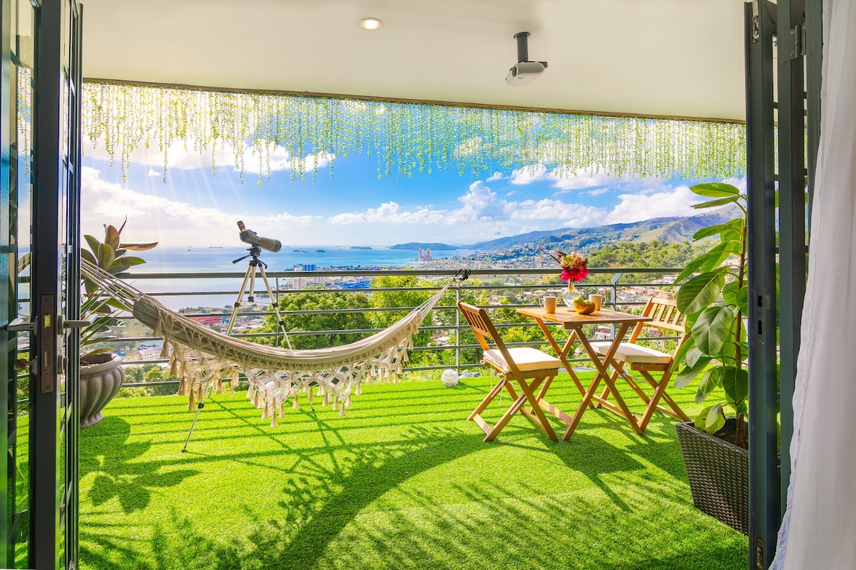 A serene balcony features a hammock and a small dining set with two chairs, all placed on lush green turf. A telescope is positioned for panoramic views of the ocean and surrounding landscape, complemented by bright sunlight streaming in.