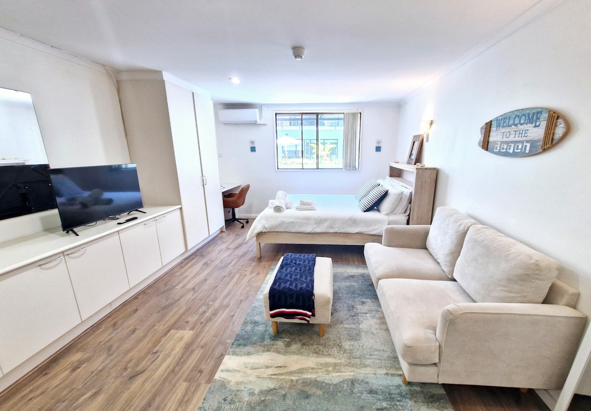 An open studio layout features a queen size bed and a two-seater sofa, complemented by a wooden desk and chair. Natural light enters through large windows, while modern furnishings are arranged on a soft area rug. A mounted TV and built-in wardrobe are positioned against a white wall.