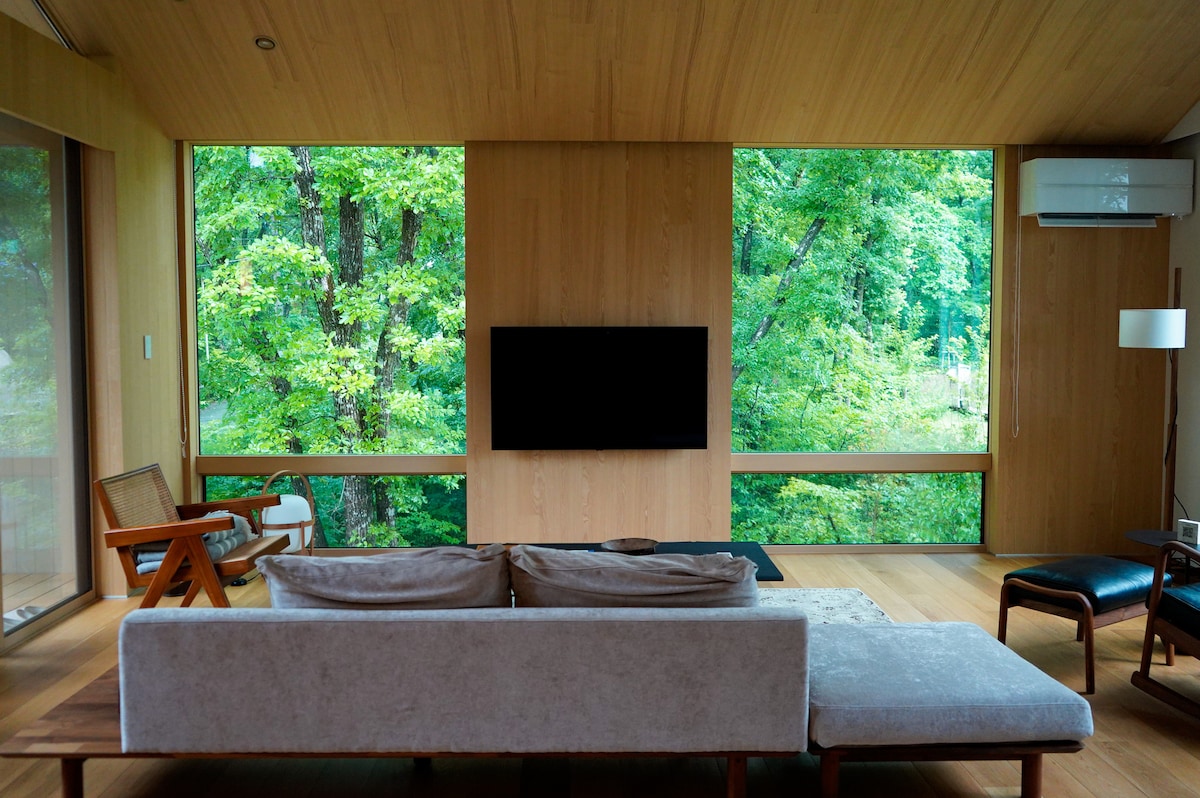 A spacious living area features large windows that provide a forest view, allowing natural light to brighten the space. A comfortable sofa and chair are arranged for relaxation, while a television is mounted on the wooden wall, creating a seamless connection with the surroundings.